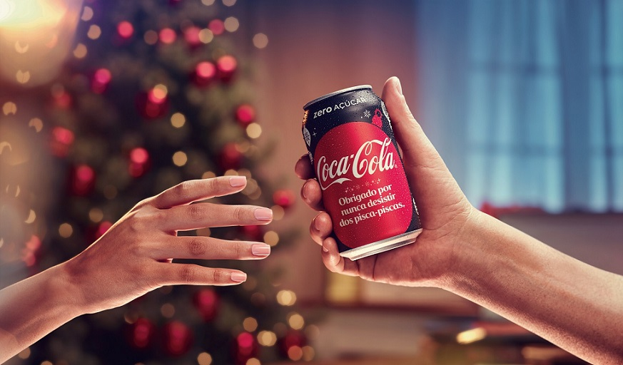 Uma mão de pessoa entregando uma lata de Coca-Cola para a outra, com um fundo de natal. Na lata há uma referência a natal